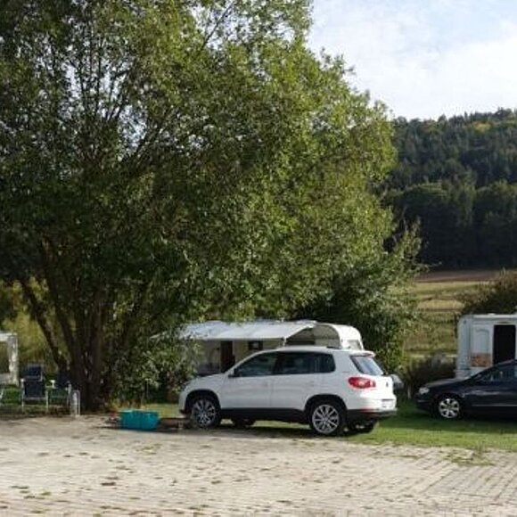 Auf dem Campingplatz stehen zwei Wohnmobile, und zwei Fahrzeuge. Ein großer Baum spendet Schatten.
