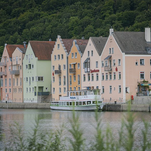 Riedenburg - Main-Donau-Kanal Der Main-Donau-Kanal im Vordergrund mit grünen Gräsern. Dahinter hat ein Personenschiff an der Schiffsanlegestelle Riedenburg angelegt. Die bekannte Promenade mit ihren Häusern ist ebenfalls zu sehen. Eingerahmt vom grün der Wälder.