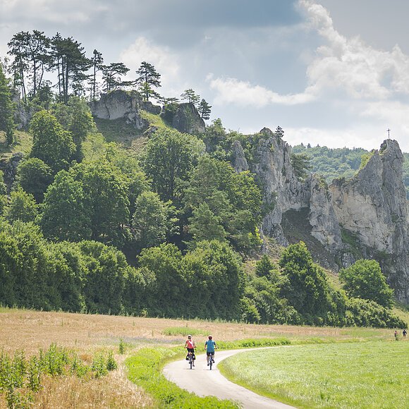 Radfahrer auf dem Altmühltal-Radweg beim Burgsteinfelsen Zwei Radfahrer auf einem Weg vor bewaldeten Felsen mit einem Kreuz auf dem höchsten Felsen.