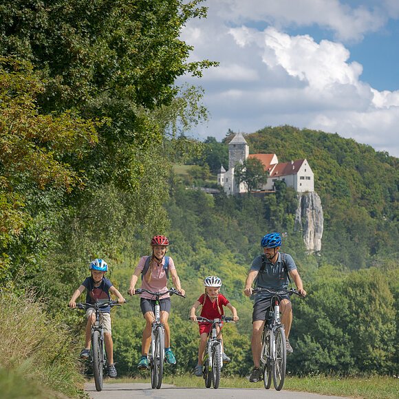 Altmühltal-Radweg (Prunn) Familie mit zwei Kindern fährt auf Fahrrädern auf einem Weg vor bewaldetem Hügel mit Burg im Hintergrund.