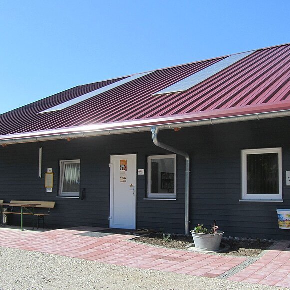 Eingang eines Gebäudes mit rotem Metalldach, zwei weißen Türen, Fenstern und einer Fahne im Hintergrund.