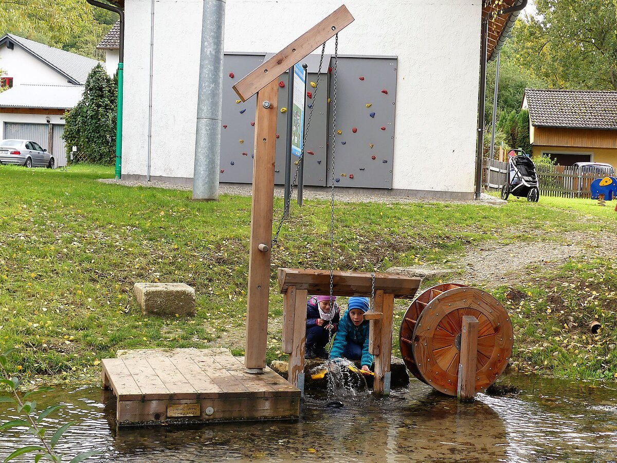 Mühlrad aus Holz mit Schwengelpumpe am Mühlbach und zwei spielende Kinder