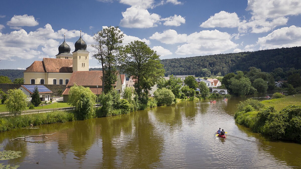 Ein Kanu rudert auf der Altmühl entlang. Links liegt Kottingwörth mit seiner Kirche St. Vitus. Die Flußufer sind saftig grün und im Hintergrund sind die bewaldeten Hänge zu sehen.