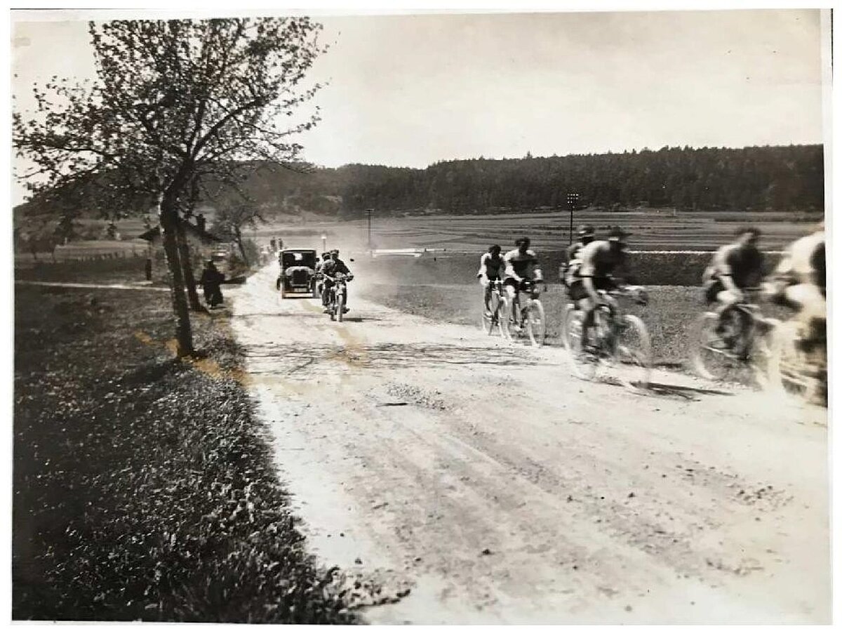 Radfahrer um 1900 fahren auf diesem Schwarzweiß-Foto auf einem Schotterweg - im Hintergrund nähert sich ein Auto ...